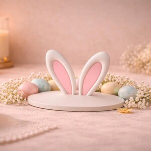 Peut inclure: Décoration d'oreilles de lapin de Pâques blanches avec l'intérieur rose, entourée d'œufs aux couleurs pastel et de fleurs blanches. Des bougies allumées dans des supports en verre sont à l'arrière-plan, créant une ambiance douce et chaleureuse. La scène se déroule sur une surface rose.