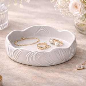 May include: A white ceramic jewelry tray with a wavy design holds a gold bracelet and rings. A gold necklace with a heart pendant and a small jewelry box are also visible. Soft lighting and flowers create an elegant setting.