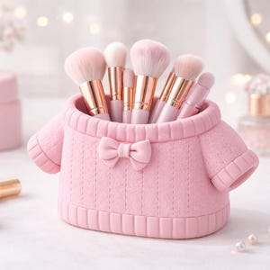 Könnte beinhalten: Ein rosa Make-up-Pinselhalter in Form eines Pullovers, verziert mit einer Schleife. Er enthält mehrere Make-up-Pinsel mit rosa Borsten und goldfarbenen Griffen. Der Hintergrund umfasst weitere Schönheitsprodukte und sanfte Beleuchtung.