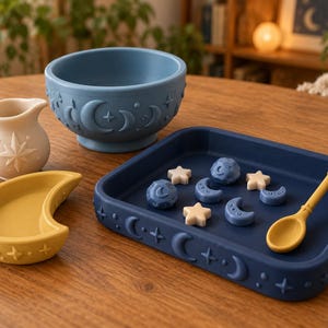 Puede incluir: Un juego de vajilla de silicona que incluye un cuenco azul, una bandeja cuadrada azul oscuro, un plato amarillo en forma de media luna, una jarra color crema y una cuchara amarilla. El cuenco y la bandeja tienen relieves con motivos de lunas y estrellas.