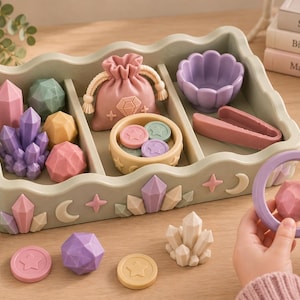 May include: A sage green divided tray decorated with pastel crystal and moon motifs, containing a pink drawstring pouch, a purple scalloped bowl, tongs, and various geometric shapes, coins, and crystal clusters in muted colors.