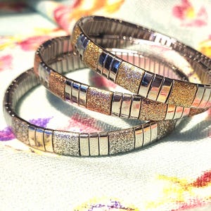 May include: Three silver and gold stretch bracelets. Each bracelet has alternating sections of silver metal and textured gold or silver glitter. The bracelets are stacked on top of each other, creating a layered effect.