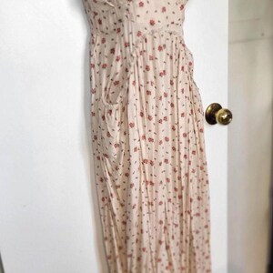 K&ouml;nnte beinhalten: Ein cremefarbenes Maxikleid mit floralem Print und Spaghettitr&auml;gern. Das Kleid hat ein figurbetontes Oberteil, eine geraffte Taille und einen flie&szlig;enden Rock mit Taschen. Kleine rote Blumen sind &uuml;ber den Stoff verstreut.