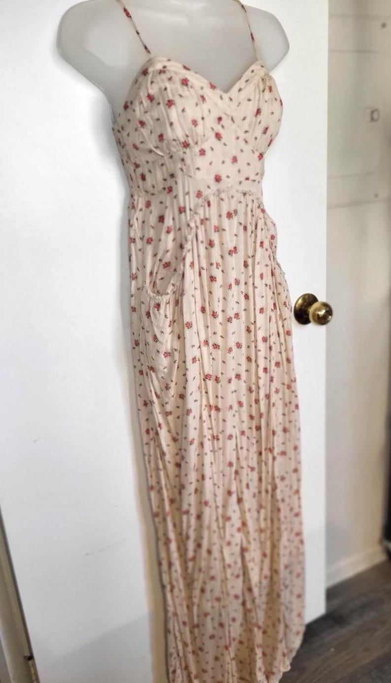 K&ouml;nnte beinhalten: Ein cremefarbenes Maxikleid mit floralem Print und Spaghettitr&auml;gern. Das Kleid hat ein figurbetontes Oberteil, eine geraffte Taille und einen flie&szlig;enden Rock mit Taschen. Kleine rote Blumen sind &uuml;ber den Stoff verstreut.