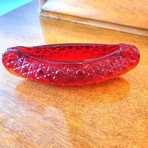 May include: A vibrant red glass dish shaped like a canoe, featuring a textured starburst and diamond pattern across its exterior surface. The translucent glass vessel has an elongated, curved form with raised edges and a polished, reflective finish.