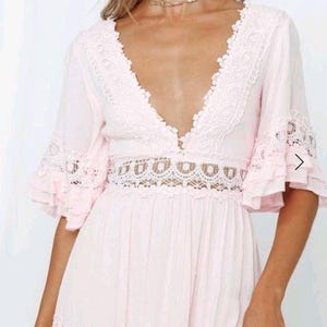 May include: A pale pink dress with a plunging V-neck and lace detailing. The dress has short, flared sleeves with lace trim and a cut-out lace design at the waist. The skirt is flowing, creating a romantic and feminine silhouette.