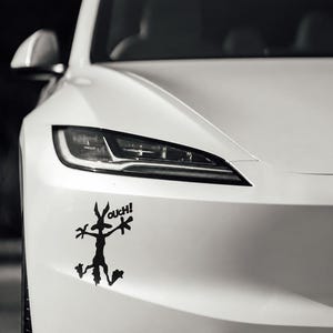 May include: A white car features a black vinyl decal on the front bumper. The decal depicts a flattened cartoon coyote character with splayed limbs and the text OUCH! positioned near the head, creating a humorous visual effect on the vehicle.