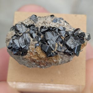 Może przedstawiać: Gromada błyszczących, czarnych kryształów, prawdopodobnie okaz mineralny, spoczywa na małej, jasnobrązowej, prostokątnej podstawie. Kryształy mają geometryczną, kątową strukturę, kontrastującą z szorstką, teksturowaną matrycą.
