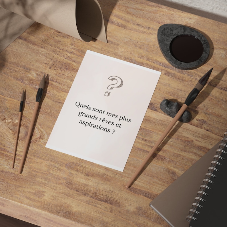 May include: A white card with a question mark and the text 'Quels sont mes plus grands rêves et aspirations ?' on a wooden surface with two paintbrushes and a black inkwell.