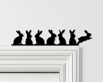 May include: A black metal decorative piece featuring a row of stylized rabbit silhouettes. Six rabbits are depicted, with one leaping off the edge. The piece is designed to sit on a ledge or trim, adding a touch of whimsy.