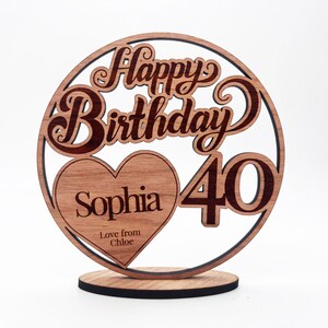 Peut inclure: Un cake topper d'anniversaire en bois avec l'inscription "Happy Birthday" dans une police décorative. Le chiffre "40" est affiché à côté d'un cœur avec le nom "Sophia" et le message "Love from Chloe". Le topper est sur une base ronde.