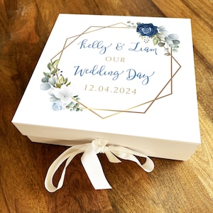 May include: A white wedding gift box with a gold geometric design and floral accents. The box features the names "Kelly & Liam" and the date "12.04.2024" in blue script. A cream ribbon is tied in a bow.