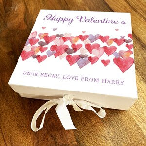 Könnte beinhalten: Weiße Geschenkbox mit einem Aquarelldesign aus rosa, roten und violetten Herzen. Die Box hat ein weißes Band und den Text "Happy Valentine's" und "Dear Becky, Love from Harry" auf der Vorderseite.