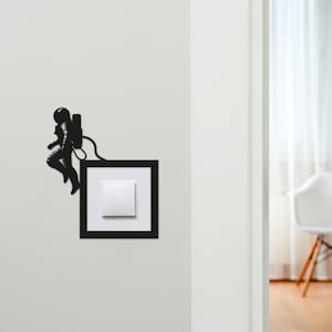 May include: A black astronaut silhouette appears to be floating next to a white light switch set in a black frame. The astronaut is wearing a spacesuit and backpack. The light switch is mounted on a white wall.