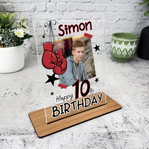 Può includere: Una cornice per foto in acrilico trasparente con una base in legno. La cornice presenta una grafica di guantoni da boxe rossi, una foto di un ragazzo e il testo "Simon Happy 10th Birthday".