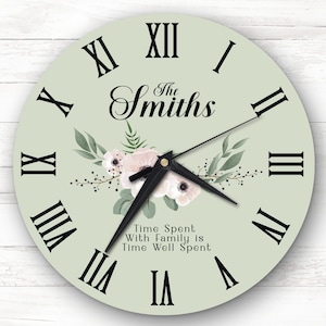 May include: A light green round clock with black Roman numeral markings and black hands. The clock face features a floral design with white flowers and green leaves. The text "The Smiths" and "Time Spent With Family is Time Well Spent" is printed on the clock face.