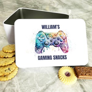 Op de afbeelding: Witte rechthoekige blik met een kleurrijk aquarelontwerp van een videogamecontroller. De tekst "WILLIAM'S GAMING SNACKS" staat op de blik gedrukt.