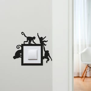 Op de afbeelding: Zwarte metalen lichtschakelaar cover met een vierkant frame en apen silhouetten. Het ontwerp toont vier apen die rond een witte lichtschakelaar klimmen. De cover is gemonteerd op een witte muur en is decoratief.