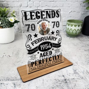 May include: A clear acrylic plaque with a black and white design that reads "Legends were born in 70 5 70 February 1954 Genuine One of a Kind Aged Perfectly All Original Parts". The plaque has a photo of a man in the center and is on a wooden stand.