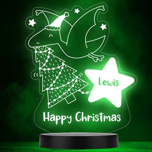 May include: A white light-up Christmas decoration with a cartoon dinosaur wearing a Santa hat and holding a Christmas tree with lights. The dinosaur is flying and there is a star with the name "Lewis" on it. The text "Happy Christmas" is at the bottom of the decoration.