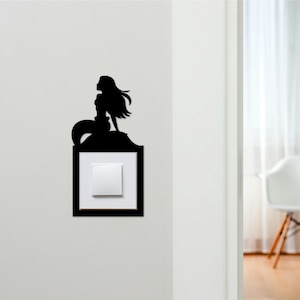 May include: Black silhouette of a mermaid perched above a light switch. The mermaid has long flowing hair and a fishtail. The light switch is white and square, set within a black rectangular frame. The design is mounted on a white wall.