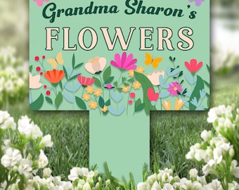 Può includere: Un paletto da giardino verde con il testo "Grandma Sharon's Flowers" in bianco. Il paletto è decorato con fiori e farfalle colorati.