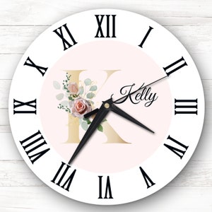 Könnte beinhalten: Eine weiße Uhr mit schwarzen römischen Ziffern für die Stunden und schwarzen Zeigern. Das Zifferblatt der Uhr ist rosa mit einem goldenen Buchstaben "K" und einem floralen Design. Der Name "Kelly" ist in Kursivschrift unter dem Buchstaben geschrieben.