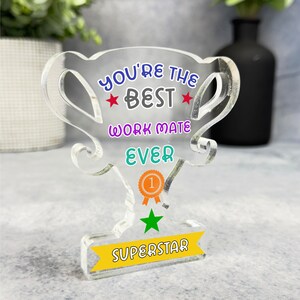 Op de afbeelding: Een transparante acryl trofee met de tekst "You're the Best Work Mate Ever" in kleurrijke letters. De trofee heeft een gouden ster en een geel lint met het nummer "1" erop. De voet van de trofee heeft een geel banner met het woord "Superstar" erop.
