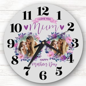 Puede incluir: Reloj redondo blanco con números y manecillas negros. El reloj tiene un diseño floral con flores moradas y una foto de dos mujeres. El texto "I love you Mum" y "Happy Mother's Day" está impreso en el reloj.