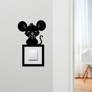 May include: A black mouse-shaped light switch cover. The mouse has large ears, a small tail, and is positioned above a white light switch set in a black frame. The design is a decorative element for a wall.
