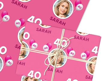May include: Pink wrapping paper with a repeating pattern of a woman's photo, the name "SARAH", the number "40", and balloons. The wrapped gift is tied with twine.