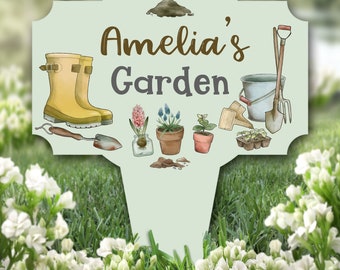 Può includere: Un cartello da giardino verde con il testo "Amelia's Garden" in lettere marroni e grigie. Il cartello presenta una varietà di attrezzi da giardinaggio e piante, tra cui un paio di stivali di gomma gialli, un annaffiatoio, una pala, una zappa e una pianta in vaso.