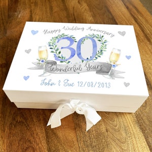 Puede incluir: Caja de regalo blanca con un diseño azul y gris. La caja tiene una cinta atada alrededor. El diseño presenta una corona con el número 30 en el centro, dos copas de champán y el texto "Happy Wedding Anniversary 30 Wonderful Years John & Sue 12/08/2013".