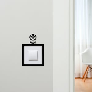 May include: A decorative light switch cover featuring a black frame with a white switch and a daisy design above. The daisy is black and adds a whimsical touch to the wall decor. The switch is centered within the frame.