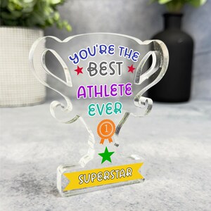 May include: A clear acrylic trophy with the text "You're the Best Athlete Ever" in colorful letters. The trophy has a gold star and a yellow number one on it. The base of the trophy has the word "Superstar" on it.