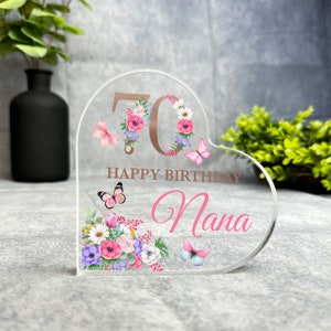 May include: A clear heart-shaped acrylic plaque with a floral design and butterflies. The number 70 is in rose gold with the text "Happy Birthday Nana".