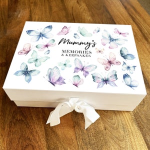 Puede incluir: Una caja de recuerdos blanca con una tapa decorada con mariposas de acuarela en tonos azules, morados y rosas. La caja está atada con una cinta blanca y presenta el texto "Mummy's Memories & Keepsakes".