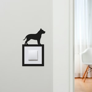 May include: A black silhouette of a dog stands atop a black square frame surrounding a white light switch. The dog is a stylized representation, likely a breed like a pit bull. The design is mounted on a white wall.