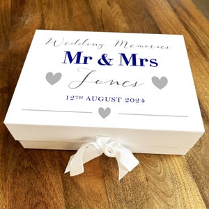 May include: White keepsake box with a blue ribbon and the text "Wedding Memories Mr & Mrs Jones 12th August 2024" with two gray hearts on each side of the text.