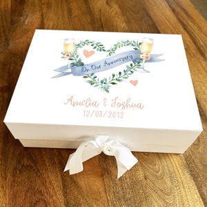 Könnte beinhalten: Weiße Geschenkbox mit einem blauen und grünen Herz-Kranz-Design, zwei Champagnergläsern und einem blauen Band mit dem Text "On Our Anniversary". Die Box ist mit einem weißen Band gebunden und hat die Namen "Amelia & Joshua" und das Datum "12/08/2012" auf der Vorderseite.