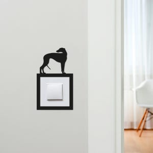 May include: A black silhouette of a dog, likely a Greyhound or similar breed, perched atop a light switch. The switch is set in a black frame against a white wall. The dog's form is sleek and elegant.