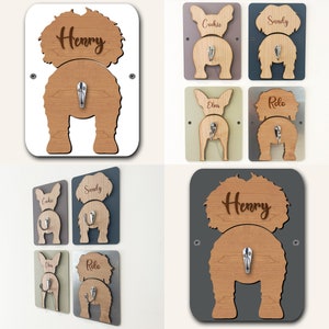 Op de afbeelding: Vier houten haken in de vorm van een hond met gepersonaliseerde namen. De haken zijn in verschillende kleuren en hebben een haak aan de achterkant om items aan op te hangen. De namen op de haken zijn "Henry", "Cookie", "Sandy", "Elvis" en "Rolo".