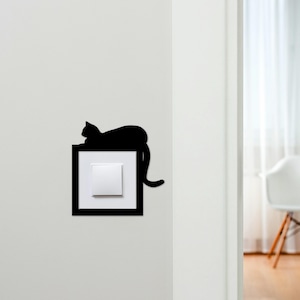 May include: A black cat-shaped light switch cover. The cat is lying on top of a square frame surrounding a white light switch. The cat's tail extends down the side. The cover is mounted on a white wall.