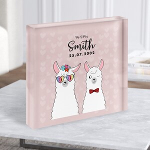 May include: A pink glass block with a white background and a heart pattern. Two white llamas are on the block, one wearing rainbow sunglasses and a flower crown, the other wearing a red bow tie. The text "Mr. & Mrs. Smith 25.07.2002" is on the block.