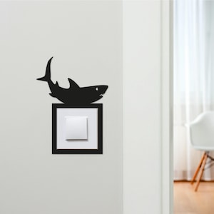 May include: A black shark-shaped light switch cover is mounted on a white wall. The shark is positioned above a white light switch within a black square frame. The design adds a playful touch to a functional item.