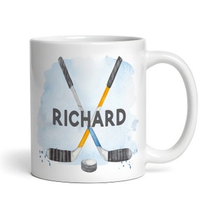 Può includere: Tazza in ceramica bianca con un design ad acquerello di due bastoni da hockey incrociati. I bastoni sono gialli, blu e neri. Il nome "RICHARD" è scritto in lettere nere sotto i bastoni. Un piccolo disco da hockey nero si trova al centro inferiore del design.