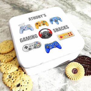 Op de afbeelding: Witte vierkante blik met een gepersonaliseerde naam "ATHONY'S" en de woorden "GAMING SNACKS". De blik is versierd met kleurrijke cartoonafbeeldingen van videogamecontrollers.