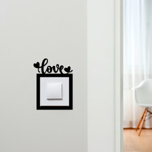 May include: A black decorative light switch cover with the word "love" in cursive script above a square frame. The word "love" has two heart shapes. The light switch is white.