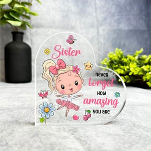 Può includere: Una placca acrilica a forma di cuore con una ballerina di cartoni animati rosa e bianca e il testo "Sorella non dimenticare mai quanto sei fantastica".