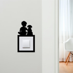 May include: Black silhouette of a child and dog sitting atop a light switch. The light switch is white and square, set within a black frame. The image is a decorative wall decal, likely for a child's room or a pet lover's space.